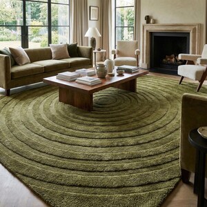 May include: A living room featuring a large, olive green rug with concentric circle design. The rug is the focal point, complemented by a brown wooden coffee table, a green sofa, and neutral-toned armchairs. The room has large windows and a fireplace.