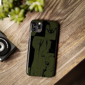 May include: A green and black phone case featuring an abstract face design. The case has a matte finish and a raised edge to protect the screen. The design includes an eye, a mouth, and other facial features.