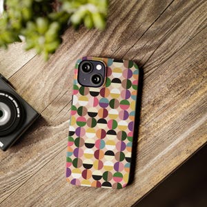 May include: A colorful phone case with a geometric pattern of half-circles in various colors, including pink, green, yellow, and purple. The case is on a wooden surface, with a camera and plant in the background.