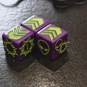 May include: Three purple dice with neon green symbols. Each die features different symbols, including arrows, alien heads, and a burst with an exclamation mark. The dice are likely for a tabletop game.