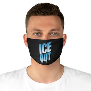 May include: Black face mask with the words "ICE OUT" in a blue and white gradient font. The text has a dripping ice effect. The mask is worn by a person with fair skin and light-colored eyes.