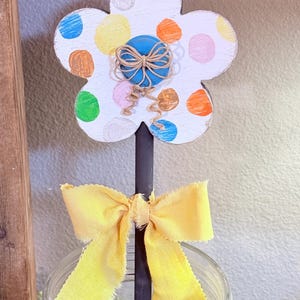 May include: A wooden flower decoration with a white base and colorful polka dots. A blue sphere is at the center, wrapped with twine. A yellow ribbon is tied below the flower. The flower is on a black rod.