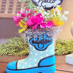May include: A decorative blue and white polka-dotted rain boot vase filled with colorful flowers and greenery. A small chalkboard sign with the word "Hello" is placed in the arrangement. The boot is sitting on a wooden surface.