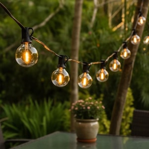 May include: String lights with clear, round bulbs illuminate a backyard setting. The warm glow of the bulbs contrasts with the green foliage in the background. A table and potted plant are in the foreground.