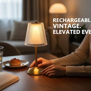 Op de afbeelding: Een kleine, goudkleurige tafellamp met een heldere, geribbelde lampenkap. De lamp staat op een houten tafel, met de tekst "RECHARGEABLE. VINTAGE. ELEVATED EVERYDAY." Een glas water, een gebakje en een open boek staan ook op de tafel.