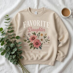 May include: A beige sweatshirt with the words "FAVORITE Daughter" in white and pink script. Below the text is a floral design with pink and white flowers. The words "EST." and "ALWAYS" flank the floral design. The words "Loved Strong Enough" are below the floral design.