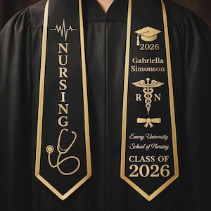 May include: Black graduation sash with gold text and accents. The left sash reads "NURSING" with a stethoscope and heartbeat line graphic. The right sash features a graduation cap, the year "2026", a name, and the Emory University School of Nursing logo.