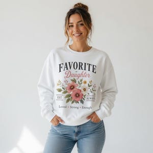 May include: White sweatshirt with the text "FAVORITE DAUGHTER" in black and pink, with a floral design. The floral design includes pink and white flowers, green leaves, and the words "Loved, Strong, Enough".