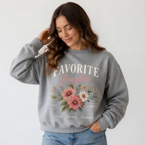 May include: A gray sweatshirt with the words "FAVORITE Daughter" in white and pink script, with a floral design below. The design includes pink, white, and yellow flowers with green leaves. The words "EST. ALWAYS Loved Strong Enough" are below the floral design.