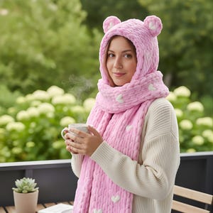 Puede incluir: Un conjunto rosa de gorro y bufanda de punto con detalles de corazones blancos. El gorro tiene orejas de oso. La bufanda está envuelta alrededor del cuello y los hombros. La persona sostiene una taza blanca con una bebida caliente.