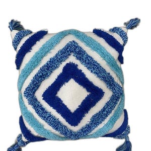 May include: Square cushion with a white centre and a geometric pattern in shades of blue. The design features a diamond shape and diagonal stripes. Each corner has a blue tassel.