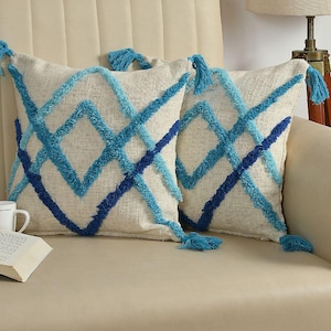 May include: Two cream-coloured textured throw cushions with a geometric design in shades of blue. Each cushion has tassels at the corners. The cushions are placed on a beige chair, creating a cosy aesthetic.