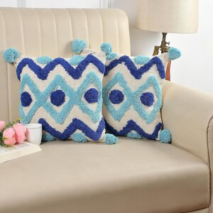 May include: Two cushions with a geometric pattern in shades of blue and light blue. The cushions have a textured design and are adorned with blue pom-poms at each corner. The cushions are on a beige sofa.