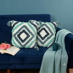 May include: Two square scatter cushions featuring a white base with a tufted diamond pattern in shades of teal and dark blue, accented with corner tassels. The cushions are displayed alongside an open book, a pink flower, and a teal knit throw blanket.