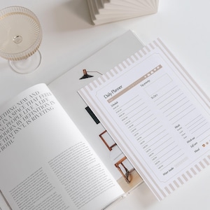 Può includere: Una composizione piatta con un'agenda giornaliera, una rivista aperta e un bicchiere di champagne. L'agenda ha un bordo a righe beige e bianco e le parole "Daily Planner". La rivista ha del testo sulla pagina.