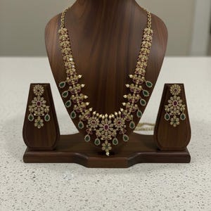 May include: A gold necklace and earring set displayed on a wooden bust. The necklace features a floral design with red, green, and clear gemstones. The earrings mirror the necklace's design, creating a matching jewellery set.