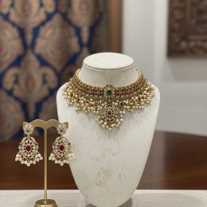 May include: A gold necklace and earring set on a white display bust and stand. The necklace features red and green gemstones, white pearls, and a central green stone. The earrings echo the necklace design, with a gold base and similar gemstones and pearls.