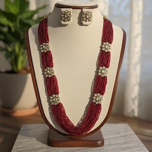 May include: A red beaded necklace and matching earrings set displayed on a mannequin. The necklace features multiple strands of small red beads, interspersed with ornate, gold-colored floral accents. The earrings have a similar design. The jewelry is presented on a cream-colored bust.