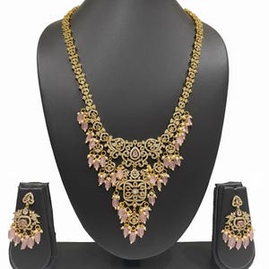 May include: A gold-toned necklace and earring set displayed on a black stand. The necklace features an ornate design with clear and pink accents, and pink teardrop-shaped beads. The matching earrings have a similar design.