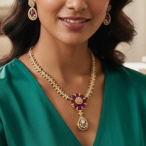 May include: A woman wearing a gold necklace and matching earrings. The necklace features a floral pendant with red gemstones and a teardrop-shaped charm. The earrings also have red gemstones. The woman is wearing a green top.
