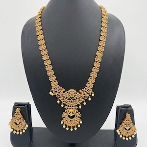 Puede incluir: Un conjunto de collar y pendientes en tono dorado con intrincado trabajo de metal en filigrana, incrustado con pequeñas piedras blancas y detalles rojos. El collar tiene un colgante central en forma de media luna con cuentas blancas.