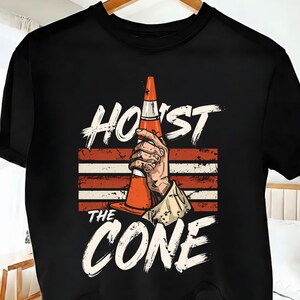 Puede incluir: Camiseta negra con un gr&aacute;fico de una mano sosteniendo un cono de tr&aacute;fico naranja. El texto "HOIST THE CONE" est&aacute; impreso encima y debajo de la imagen en una fuente blanca desgastada, con rayas rojas y blancas en el fondo.
