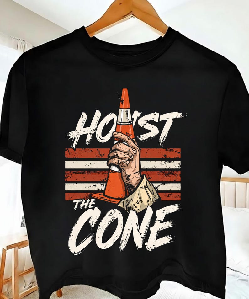 Puede incluir: Camiseta negra con un gr&aacute;fico de una mano sosteniendo un cono de tr&aacute;fico naranja. El texto "HOIST THE CONE" est&aacute; impreso encima y debajo de la imagen en una fuente blanca desgastada, con rayas rojas y blancas en el fondo.