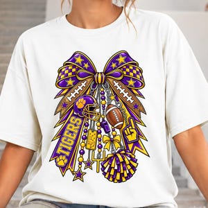 Puede incluir: Una camiseta color crema con un gran gráfico de un lazo morado y dorado adornado con elementos de fútbol americano, incluyendo un casco, un balón, un silbato, un pompón, un poste de meta y la palabra TIGERS impresa en una cinta.