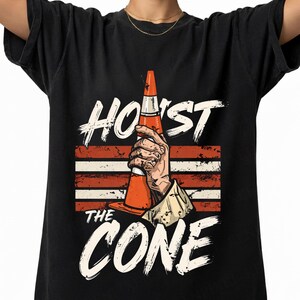 Puede incluir: Camiseta negra con las palabras blancas "HOIST THE CONE". El dise&ntilde;o presenta una mano sosteniendo un cono de tr&aacute;fico naranja sobre un fondo de rayas rojas y blancas.