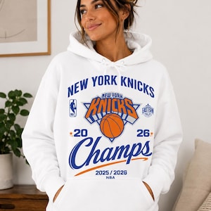 May include: A white hooded sweatshirt featuring a blue and orange graphic on the front. The text reads New York Knicks, 2025/2026 NBA, and Champs. The design includes the New York Knicks logo, a basketball, and the NBA logo in blue and orange ink.