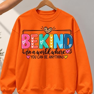 May include: A bright orange crewneck sweatshirt featuring a graphic print with the text BEKIND in colourful block letters, a leopard print heart, and the phrase in a world where YOU CAN BE ANYTHING with small heart icons.