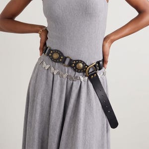May include: A black leather belt featuring brass-toned circular studs and hardware, paired with a silver-toned chain belt composed of interconnected butterfly charms. The black belt has a wide strap with a buckle and a long, dangling end piece.