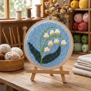 Op de afbeelding: Een blauw geborduurd rond stuk met een lelietjes-van-dalen ontwerp, gepresenteerd op een kleine houten ezel. De afbeelding bevat knutselbenodigdheden zoals garen en houten spoelen, wat wijst op een knutselthema. De borduurwerk is in een ronde lijst.