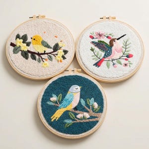 May include: Three embroidered bird artworks in wooden hoops. One shows a yellow bird on a branch with yellow flowers, another a hummingbird with pink and teal feathers, and the last a blue and yellow bird on a branch with white flowers.