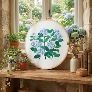Könnte beinhalten: Ein runder Stickrahmen mit einem detaillierten floralen Design aus blauen Hortensien mit grünen Blättern. Die Stickerei befindet sich auf weißem Stoff, umrahmt von einem natürlichen Holzrahmen. Das Kunstwerk steht auf einer Holzoberfläche.