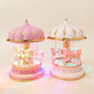 May include: Two carousel music boxes, one with a pink canopy and base, the other with a white canopy and base. Both feature gold accents, a central gold pole, and multiple miniature horses with gold saddles, illuminated by internal LED lights.