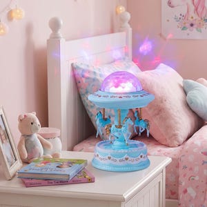 May include: A light blue carousel lamp featuring a rotating top with a glowing purple dome light and three small blue horses. The base is decorated with scalloped edges and heart patterns, emitting a soft, colorful light effect.