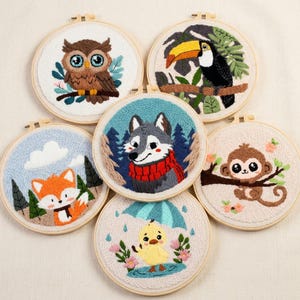 May include: Six embroidered hoops showcasing animal designs. The collection includes an owl, toucan, wolf, fox, monkey, and duck. Each animal is depicted with its surroundings, like trees or rain, and created using colourful thread.