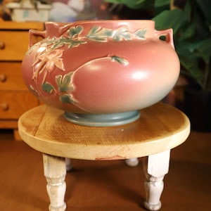 May include: A decorative, round, pink ceramic vase with green and pink floral relief sits on a small, round wooden stool with white legs. The vase has two handles and a light blue base.