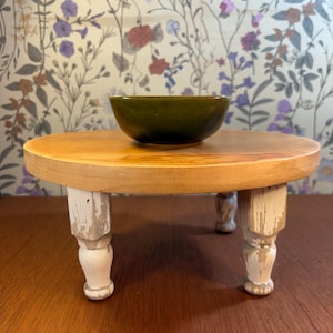 May include: A small, round wooden plant stand with a light brown top and four white, distressed legs. A dark green ceramic bowl sits on top. The stand is on a dark brown surface, with a floral wallpaper background.