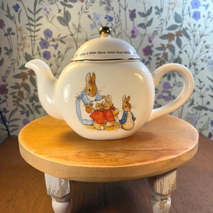 May include: A white ceramic teapot with a gold-colored knob on the lid. The teapot features illustrations of rabbits in clothing. The lid has the text "...upon a time there were four little..." The teapot sits on a small, round wooden stand with white distressed legs.
