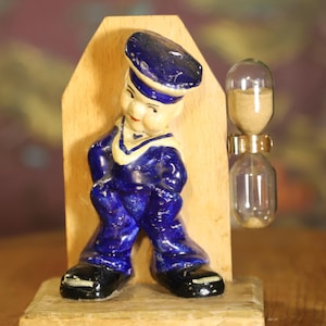May include: A vintage sailor figurine in a blue uniform with a white collar and black shoes. The figurine is attached to a wooden base with an hourglass. The hourglass is made of glass and contains sand.