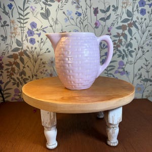 May include: A light purple ceramic pitcher with a woven texture sits on a small, round wooden pedestal with white, distressed legs. The background features a floral wallpaper.