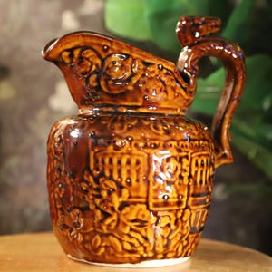 May include: A brown ceramic pitcher with a glossy finish. The pitcher features ornate embossed floral and geometric patterns. It has a curved handle and a spout for pouring. The pitcher is displayed on a wooden surface.