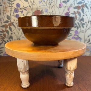May include: A brown ceramic bowl sits on a small, round wooden stand with white, distressed legs. The bowl has a glossy finish. The stand is light brown with a distressed white paint on the legs. The background features a floral wallpaper.