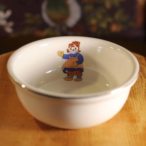 May include: A white ceramic bowl with a gold rim. The bowl features a colorful illustration of a character in a blue dress and orange apron. The bowl is sitting on a wooden surface.