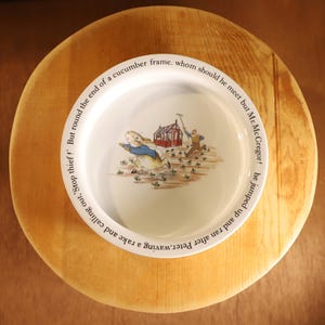 May include: A white ceramic plate featuring a colorful illustration of Peter Rabbit running from a farmer. The plate has text around the rim and is set on a wooden surface.