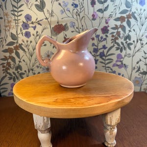 May include: A small, pink ceramic pitcher with a handle sits on a round, wooden plant stand. The stand has three short, white-painted legs with distressed detailing. The background features a floral wallpaper.