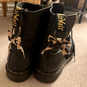 Puede incluir: Botines de cuero negro con costuras amarillas y una lengüeta con el texto "AirWair". Cada bota tiene un pequeño lazo con un estampado de vaca en blanco y negro.