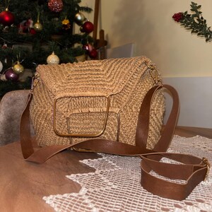 May include: A woven straw handbag with a brown leather shoulder strap. The bag has a rectangular shape with a gold-colored metal frame and clasp. The bag is sitting on a table with a white lace doily.